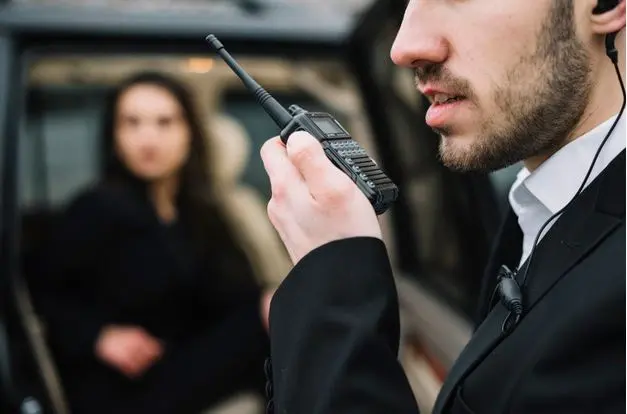 Imagem de homem branco falando em walkie talkie fazendo segurança empresarial e mulher atrás desfocada.