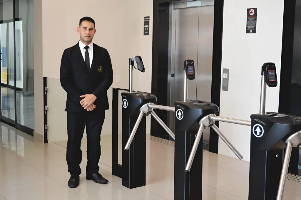Porteiro recebendo pessoas ao lado de catracas para controle de acesso de visitantes em prédio