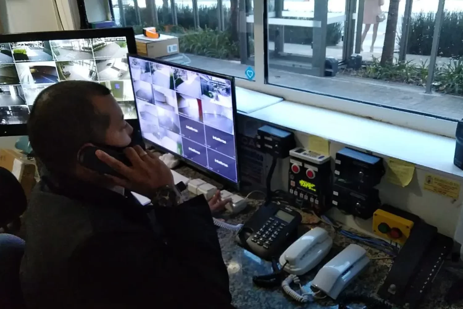 Trabalhador de portaria terceirizada falando ao telefone enquanto monitora câmeras de segurança de prédio.